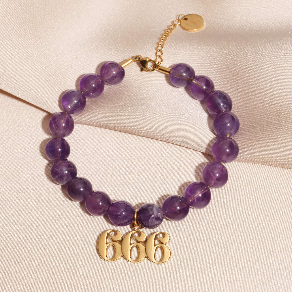 666 gold charm on beaded amethyst bracelet laying on silk