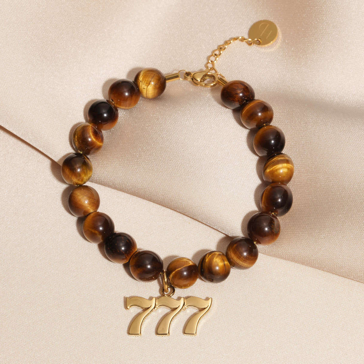 777 Gold charm on beaded Tigers Eye bracelet laying on silk
